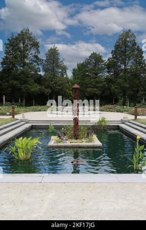 Ein künstlich anbauter Teich mit Wasserpflanzen und eine Skulptur im Botanischen Garten Boerner mit einem Rosengarten und einer Reihe dichter Bäume im Hintergrund Stockfoto