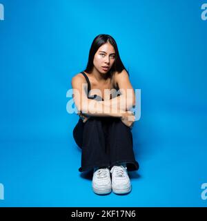 Multirassischer Teenager, der auf dem Boden in Fetalposition auf blauem Hintergrund mit Kopierraum sitzt Stockfoto