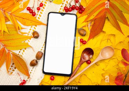 Nachbau eines Smartphone-Displays auf einem Hintergrund in gelb-orangenen Rottönen: Ein weiß gestricktes Karo hinterlässt Beeren Löffel Haselnüsse. Vorlage für den Herbst Stockfoto