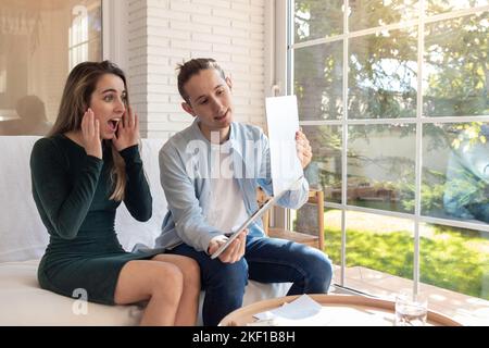 Die Frau überraschte etwas, das auf dem Laptop mit staunenden Gesichten und Händen angesehen wurde, während der Mann den Bildschirm im Wohnbereich zeigte Stockfoto