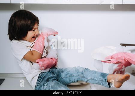 Hausfrau Mutter in rosa Handschuhen wäscht Geschirr mit ihrem Sohn von Hand in der Spüle mit Waschmittel. Ein Mädchen in Weiß und ein Kind mit einem Wurf putzen das Haus Stockfoto