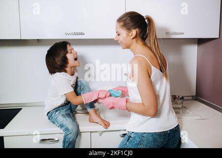 Hausfrau Mutter in rosa Handschuhen wäscht Geschirr mit ihrem Sohn von Hand in der Spüle mit Waschmittel. Ein Mädchen in Weiß und ein Kind mit einem Wurf putzen das Haus Stockfoto