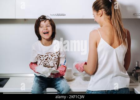 Hausfrau Mutter in rosa Handschuhen wäscht Geschirr mit ihrem Sohn von Hand in der Spüle mit Waschmittel. Ein Mädchen in Weiß und ein Kind mit einem Wurf putzen das Haus Stockfoto