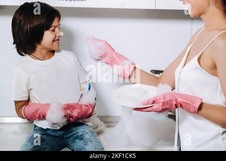 Hausfrau Mutter in rosa Handschuhen wäscht Geschirr mit ihrem Sohn von Hand in der Spüle mit Waschmittel. Ein Mädchen in Weiß und ein Kind mit einem Wurf putzen das Haus Stockfoto