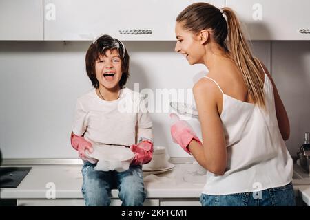 Hausfrau Mutter in rosa Handschuhen wäscht Geschirr mit ihrem Sohn von Hand in der Spüle mit Waschmittel. Ein Mädchen in Weiß und ein Kind mit einem Wurf putzen das Haus Stockfoto