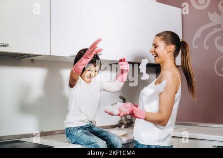 Hausfrau Mutter in rosa Handschuhen wäscht Geschirr mit ihrem Sohn von Hand in der Spüle mit Waschmittel. Ein Mädchen in Weiß und ein Kind mit einem Wurf putzen das Haus Stockfoto