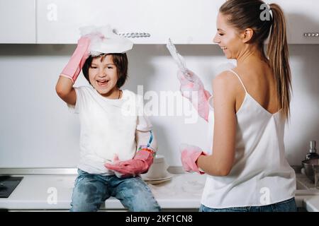 Hausfrau Mutter in rosa Handschuhen wäscht Geschirr mit ihrem Sohn von Hand in der Spüle mit Waschmittel. Ein Mädchen in Weiß und ein Kind mit einem Wurf putzen das Haus Stockfoto