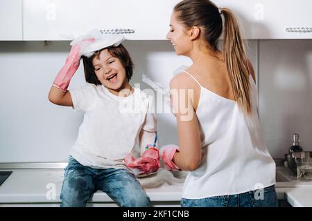 Hausfrau Mutter in rosa Handschuhen wäscht Geschirr mit ihrem Sohn von Hand in der Spüle mit Waschmittel. Ein Mädchen in Weiß und ein Kind mit einem Wurf putzen das Haus Stockfoto