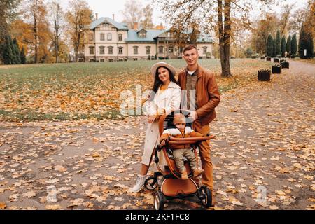 Vater und Mutter auf einem Spaziergang mit einem Kind im Kinderwagen im Herbstpark auf dem Hintergrund des Anwesens. Eine Familie spaziert durch den Goldenen Herbst Stockfoto