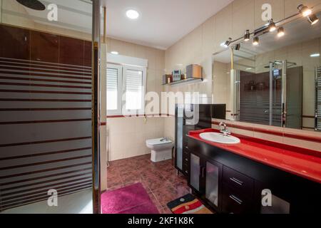 Bad mit weißen Porzellanarmaturen, roten Steinablagen, dunklen Holzschränken und großem rahmenlosen Spiegel Stockfoto