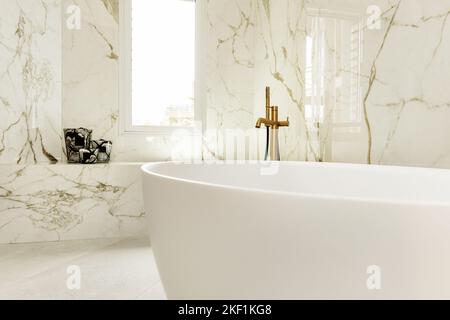 Badezimmer mit großer Badewanne mit Messingarmaturen und Marmorfliesen mit Aluminiumfenster und eingelassenem Sitz aus dem gleichen Material Stockfoto