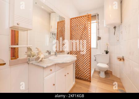 Toilette mit Spiegel an der Wand, weiße Holzmöbel mit Marmorplatte und Holzgitter in der Dusche Stockfoto