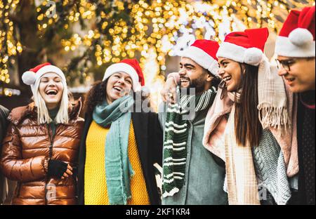 Trendige Menschengruppe, die unter Weihnachtsbaumschmuck läuft - Konzept im Winterleben mit fröhlichen Freunden, die rote weihnachtsmützen tragen und dabei Spaß am Urlaub haben Stockfoto