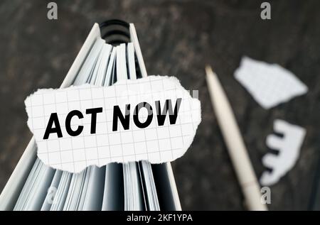 Auf dem Tisch steht ein Notizbuch, auf dem ein zerrissenes Papier mit der Aufschrift - Act Now liegt. Der Stift liegt außerhalb des Schärfebereichs. Stockfoto