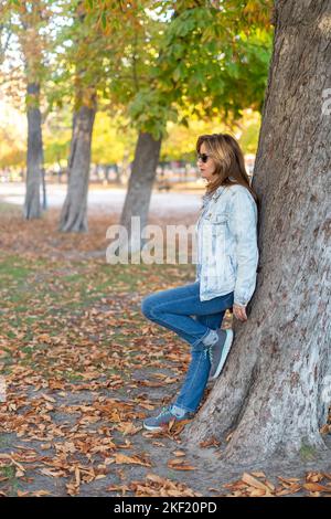 Reife weiße Frau, die sich in einem öffentlichen Park an einem großen Baum lehnt, in dem im Herbst Blätter auf den Boden fallen. Stockfoto