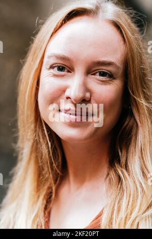 Nahaufnahme eines vertikalen Porträts einer ruhigen und attraktiven jungen blonden Frau. Sie blickt mit einem zufriedenen Lächeln auf die Kamera Stockfoto