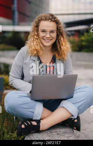 Vorderansicht einer lockigen Frau, die eine Brille trägt und die Kamera anschaut, Zeit mit ihrem Computer im Freien verbringt, aus der Ferne arbeitet oder studiert. Lächelnd Stockfoto
