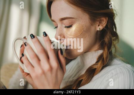 Porträt einer schönen Sommersprossen-Frau mit Augenflecken, die sich auf dem gemütlichen Sofa ausruhen und eine Tasse Tee trinken, um das Wochenende zu genießen. Unter Augenflecken. Freizeit Stockfoto