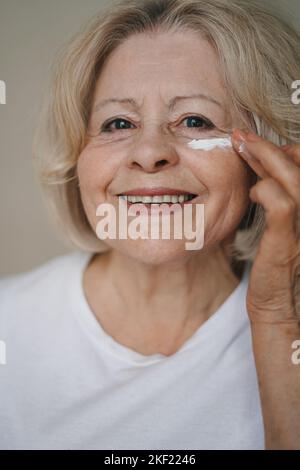 Ältere Frau, die feuchtigkeitsspendende Creme unter ihre Augen auf braunem Hintergrund auftragen. Schönheitsbehandlung mit kosmetischem Produkt. Make-up-Produkt. Dermatologie Stockfoto
