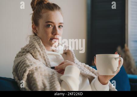 Porträt einer jungen kranken Frau, die auf die Kamera schaut, die auf dem Sofa sitzt und Tee trinkt. Tasse Tee, Kaffee und Chill. Freizeit genießen, Wochenende zu Hause Stockfoto