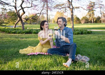 Ein Paar auf einer karierten Picknickdecke im Forest Park mit einer Flasche Wein und roten Knospenbäumen dahinter. Stockfoto