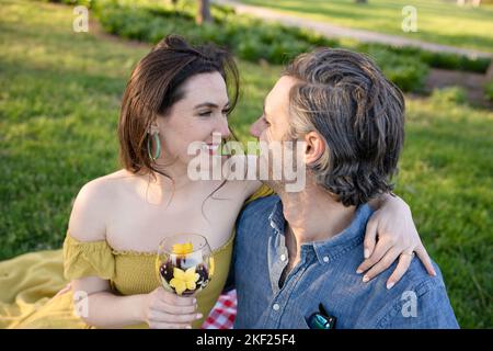 Ein Paar auf einer karierten Picknickdecke im Forest Park mit einer Flasche Wein und roten Knospenbäumen dahinter. Stockfoto