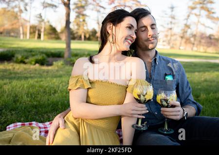 Ein Paar auf einer karierten Picknickdecke im Forest Park mit einer Flasche Wein und roten Knospenbäumen dahinter. Stockfoto