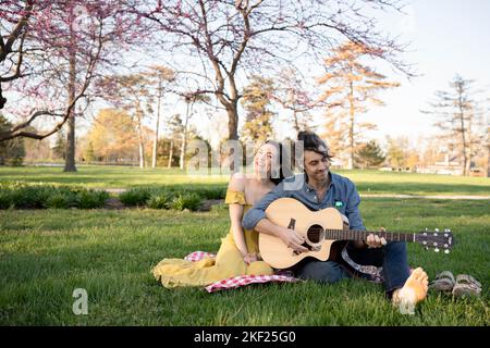 Verliebte Paare bei einem Picknick im Forest Park. Der Mann spielt eine Gitarre für seine Frau. Stockfoto