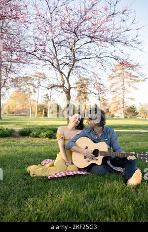 Verliebte Paare bei einem Picknick im Forest Park. Der Mann spielt eine Gitarre für seine Frau. Stockfoto