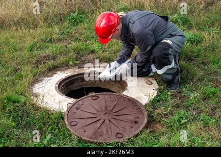 Ein männlicher Klempner in Overalls und ein Helm in der Nähe eines Wasserbochs schreibt die Messungen und die Messwerte des Wasserzählers auf. Stockfoto