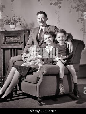 1940S GLÜCKLICH LÄCHELNDE FAMILIE VON VIER SITZEN ZUSAMMEN IM WOHNZIMMER STUHL VON KONSOLE RADIO PAPA MUTTER SOHN TOCHTER SUCHEN BEI DER KAMERA - J4984 HAR001 HARS DRINNEN NOSTALGISCHES PAAR 4 SCHRANK MÜTTER ALTE ZEIT ZUKUNFT NOSTALGIE BRUDER ALTE MODE SCHWESTER 1 JUGENDLICHE STIL JUNGE ERWACHSENE SÖHNE ERFREUT FREUDE LIFESTYLE ZUFRIEDENHEIT FRAUEN BRÜDER STUDIO SCHUSS GESUNDHEIT ZU HAUSE LEBEN KOPIEREN RAUM FREUNDSCHAFT HALBLANGE TÖCHTER FÜRSORGLICH MÄNNER GESCHWISTER SCHWESTERN VÄTER S&W BLICKKONTAKT ANZUG UND KRAWATTE KONSOLE GLÜCK FRÖHLICH WOHNZIMMER VÄTER STOLZ DURCH IN DER GESCHWISTER LÄCHELT VERBINDUNG FRÖHLICH STILVOLL UNTERSTÜTZEN ZUSAMMENARBEIT WACHSTUM Stockfoto