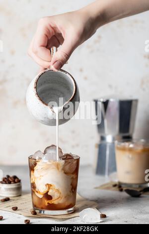 Milch in Kaffee mit Eiswürfeln mit schönen Wendungen auf hellem Hintergrund von Hand gießen. Vertikale Ausrichtung. Iced Latte Coffee Stillleben Stockfoto