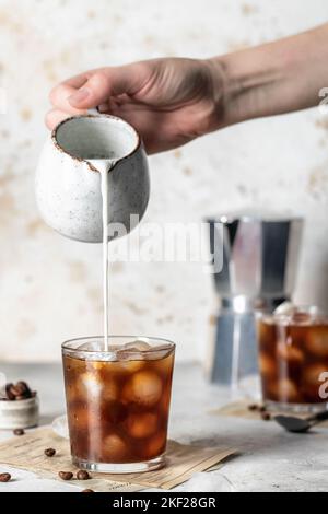 Frau gießt Milch in Kaffee mit Eiswürfeln auf hellem Hintergrund. Iced Latte Coffee Stillleben. Vertikale Ausrichtung Stockfoto