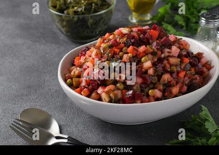 Vinaigrette-Salat aus Rüben, Kartoffeln, Kohl, grünen Erbsen, Karotten, Gurken, Zwiebeln und Pflanzenöl in einer weißen Schüssel auf grauem Hintergrund Stockfoto