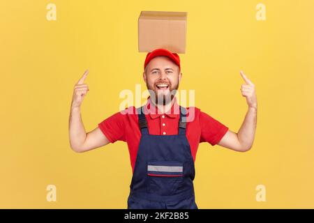 Porträt eines aufgeregten Kuriermanns in blauer Uniform, der auf einen Karton auf dem Kopf zeigt und mit fröhlichem Ausdruck auf die Kamera blickt. Innenaufnahme des Studios isoliert auf gelbem Hintergrund. Stockfoto