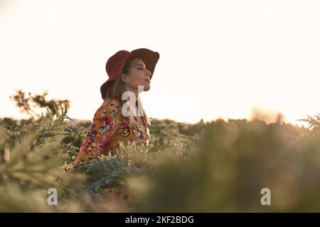 kaukasische reife Frau in einem Blumenkleid und einem roten Hut ruhig und entspannt meditieren mit geschlossenen Augen an einem sonnigen Tag Stockfoto