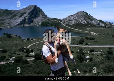 lateinischer Mann in Sonnenbrille mit einem großen Hund in den Armen in der Nähe des Sees und der Berge, covadonga asturias, spanien Stockfoto