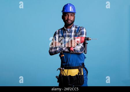 Studiofoto des Reparaturwerkers, der die Kamera ansah, mit gekreuzten Armen über seiner Brust und einem Akku-Elektrobohrer. Bauarbeiter trägt Overalls und Gürtel mit allen Arbeitswerkzeugen. Stockfoto