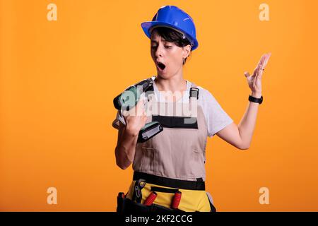Junge, fröhliche Frau mit elektrischer Nagelpistole auf gelbem Hintergrund. Weibliche Bauherrin trägt Overalls und Hardhut im Studio, mit poer Bohrwerkzeug für die Wartung. Stockfoto