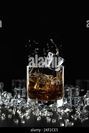 Ein Spritzer Whiskey und ein fallender Eiswürfel in einem Glas auf schwarzem Hintergrund zwischen zerkleinertem Eis, ähnlich wie Diamanten Stockfoto