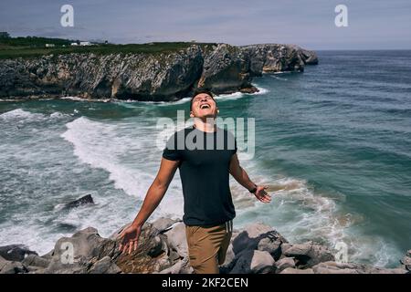 lateinischer Junge in schwarzem T-Shirt und braunen Shorts, der in den Himmel blickt und mit ausgestreckten Armen auf den Felsen in der Nähe der Meereswellen schreit Stockfoto
