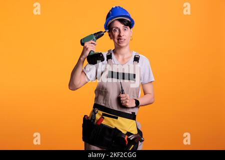 Frau Reparatur Angestellter hält Power-Bohrpistole und Spaß haben, benommen handeln. Bauarbeiterin trägt Hardhut und Umbauuniform und ist beim Renovieren der Nagelpistole lustig. Stockfoto