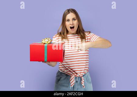 Porträt der erstaunt überrascht blonde Frau trägt gestreiftes T-Shirt halten und zeigen rot Geschenkbox, Geschenk für den Urlaub zu präsentieren. Innenaufnahme des Studios isoliert auf violettem Hintergrund. Stockfoto