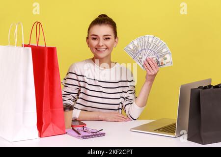 Porträt einer zufriedenen Büromanagerin, die mit Einkaufstaschen am Arbeitsplatz sitzt, einen Fan von Dollars zeigt, Cashback, die Kamera anschaut. Studio-Innenaufnahme isoliert auf gelbem Hintergrund. Stockfoto