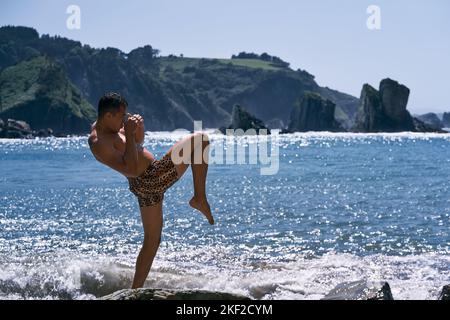 lateinischer junger Mann, der einen Tiger im Profil trägt, seine Arme gefaltet und dabei nasse Kampfkunsthaltungen mit einem Bein im Meerwasser in der Nähe des macht Stockfoto