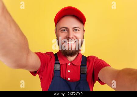 Optimistischer, positiver Arbeiter mit blauer Uniform, der Selfie macht, die Kamera mit toothy POV, Standpunkt des Fotos anschaut. Innenaufnahme des Studios isoliert auf gelbem Hintergrund. Stockfoto