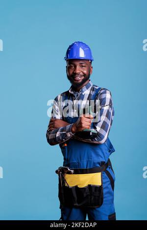 Professioneller Auftragnehmer mit Schutzhelm und Werkzeuggürtel auf hellblauem Hintergrund. afroamerikanischer Baumeister mit Akku-Bohrmaschine und zweckarmigem Taillenband, während er lächelnd auf die Kamera blickt. Stockfoto