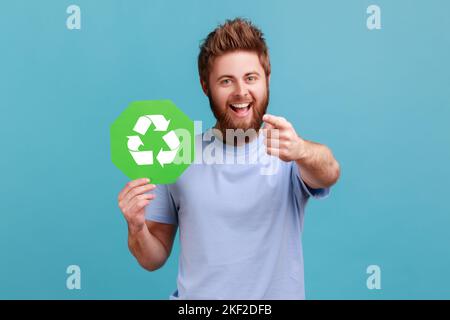 Porträt eines äußerst glücklichen, aufgeregt bärtigen Mannes mit grünem Recycling-Schild, Umweltschutz, Ökologie-Konzept, zeigt auf die Kamera. Innenaufnahme des Studios isoliert auf blauem Hintergrund. Stockfoto