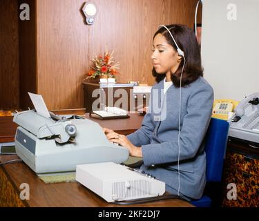 1970S AFRIKANISCH-AMERIKANISCHE FRAU IM BÜRO TIPPT AUF ELEKTRISCHER SCHREIBMASCHINE UND HÖRT DIKTAT VON EINEM DIKTIERGERÄT MIT HEADSET - KO454 HAR001 HARS HALBLANGE DAMEN PERSONEN VERTRAUEN HEADSET DIKTAPHONE FÄHIGKEIT BERUF FÄHIGKEITEN EINGABE AFROAMERIKANER AFROAMERIKANISCHE SCHWARZE ETHNIE BÜROANGESTELLTE BERUFE GAL FREITAG ADMINISTRATOR SEKRETÄRE STILVOLLE UNTERSTÜTZUNG AMANUENSIS DIKTAT JUNGE ERWACHSENE FRAU KLERIKALE HAR001 ALTMODISCHE AFROAMERIKANER Stockfoto