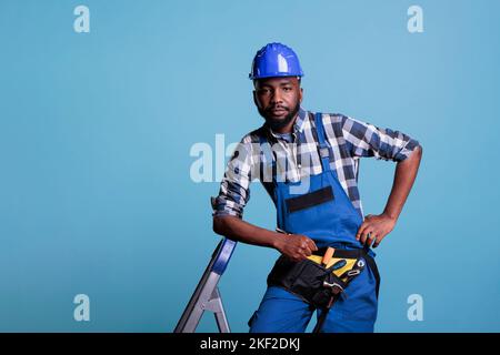 Seriöse Baumeister, die sich auf einer Leiter mit Overalls und Schutzhelm lehnen und vor hellblauem Hintergrund auf die Kamera blicken. Studioaufnahme mit Konzept der Bauindustrie. Stockfoto